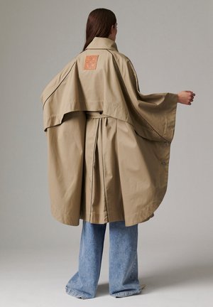 Personne portant un trench-coat beige ample avec un détail de cape dans le dos et un passepoil noir, associé à un jean bleu à jambes larges, debout devant un fond uni.