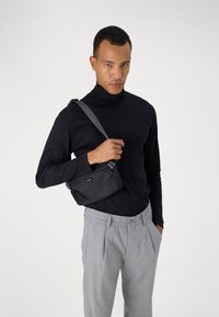 Sac noir en bandoulière avec un tissu texturé, doté d'une seule sangle réglable et d'une petite étiquette de marque, tenu par une personne portant un pull col roulé noir.