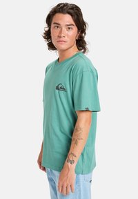 Short-sleeve, teal cotton t-shirt with a black wave logo on the left chest and a tag on the left sleeve, paired with light blue jeans.
