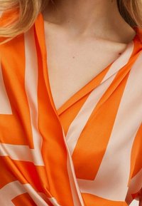 Top envolvente naranja y crema con patrones geométricos, fabricado en tejido suave, diseño de escote en V y caída suave en el cuello.
