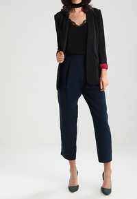 Czarna marynarka z czerwonymi akcentami na mankietach, w połączeniu z czarnym topem wykończonym koronką oraz granatowymi spodniami o zwężanym kroju. Modelka ma na sobie zielone obcasy.