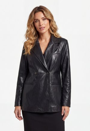 Vrouw met golvend blond haar die een zwarte, dubbelgeknoopte leren blazer draagt over een zwarte jurk, staand tegen een effen achtergrond.