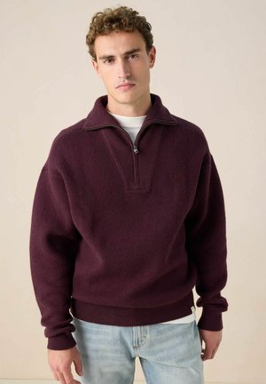 Young man with curly hair wearing a maroon quarter-zip sweater and light blue jeans, standing against a plain beige background.