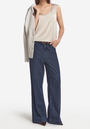 Kvinde iført cremefarvet, ærmeløs bluse, mørke, vidde jeans med brun bælte, som holder en lysebeige, struktureret jakke over skulderen.