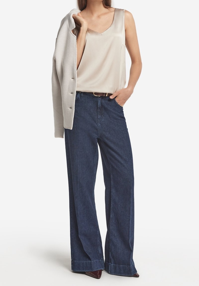 Femme portant un chemisier crème sans manches, un jean large foncé avec une ceinture marron, tenant une veste texturée beige clair sur l'épaule.