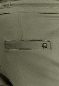 Olijfgroene broek met een zachte textuur, voorzien van een achterzak met een hexagonaal zwart en gouden accent.
