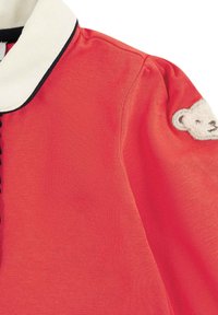 Rotes gestricktes Oberteil mit einem weißen Kragen, der mit schwarzem Besatz verziert ist, Knopfdetails und einem strukturierten Bärenpatch an den Ärmeln.