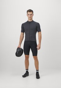 Un maillot de cyclisme ajusté, gris foncé, avec une fermeture éclair à l'avant, associé à un short noir. Le modèle tient un casque noir et porte des chaussures de cyclisme noires.