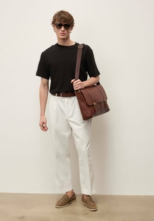 Homme portant un t-shirt noir, un pantalon blanc, une ceinture et des chaussures marron, portant un sac à bandoulière en cuir marron, debout contre un mur uni.