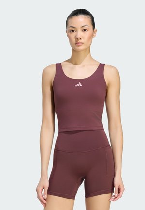 Kvinde stående vendt fremad iført matchende burgunderfarvet atletisk crop top og højtaljede shorts med et lille logo på brystet.