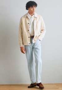 Beige Baumwolljacke mit schwarzen Knöpfen, darunter ein weißes Hemd, hellblaue Jeans und braune Schuhe. Das Model steht vor einem neutralen Hintergrund.