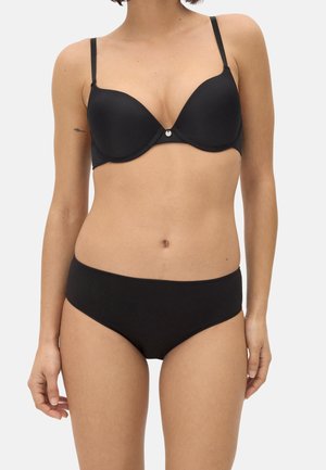 Mujer con un sujetador negro con aro y tirantes finos, y unas braguitas negras de talle medio a juego, posando frente a un fondo liso.