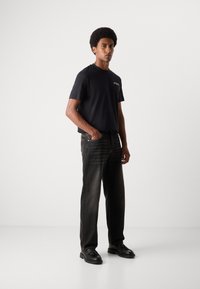 Schwarzes Baumwoll-T-Shirt mit Text-Logo, kombiniert mit dunklen Jeans im Straight-Leg-Schnitt mit verwaschenem Finish und schwarzen Schnürschuhen.