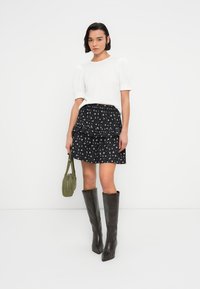 Haut blanc texturé à manches courtes, jupe noire à volants avec motif floral, sac vert olive et bottes montantes marron foncé.