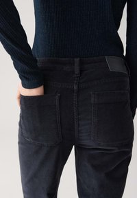 Persoon in een zwarte geribde trui en donkere corduroy broek met hand in de achterzak, weergegeven van taille tot midden boven de knie.
