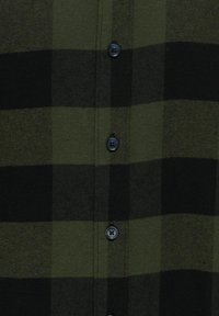 Cămașă din flanel în carouri verzi și negre, cu închidere cu butoane și o textură moale. Butoanele sunt de culoare închisă, cu un design cu patru orificii.