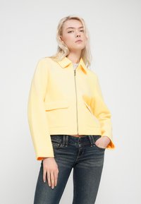 Κίτρινο cropped μπουφάν με φερμουάρ στο μπροστινό μέρος, μυτερό γιακά και δύο μπροστινές τσέπες, σε συνδυασμό με στενά σκουρό μπλε τζιν. Λεία υφή.