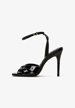 Sandalia de tacón alto de charol negro con tiras entrelazadas, punta abierta y correa ajustable en el tobillo. Diseño elegante con un acabado brillante.