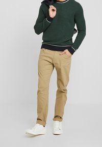 Mörkgrön texturerad tröja med vita och marinblå accenter, kombinerad med beige chinos och vita sneakers, som visar en avslappnad outfit.