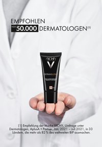 Black tube of Vichy Dermablend foundation, featuring a white and silver logo, held in a hand against a blurred white lab coat background.