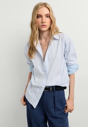 Jonge vrouw met lang blond haar draagt een lichtblauw gestreept shirt met opgerolde mouwen en donkerblauwe broek met hoge taille en een zwarte riem.