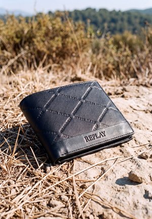 Una cartera negra plegable de Replay está sobre un terreno seco y pedregoso, entre hierba de color pajizo.