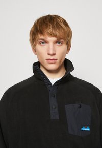 Pullover in pile nero con colletto alto, caratterizzato da una pattina dei bottoni blu navy e una tasca sul petto. Include un logo KAVU sulla tasca.