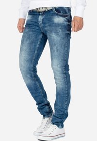 Jean en denim bleu avec une coupe slim, présentant des délavages clairs et un léger effet de décoloration. Associé à des baskets blanches et une ceinture grise.