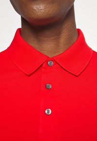 Cou du haut et poitrine d'une personne portant un polo rouge vif avec trois boutons visibles et un col rabattu.