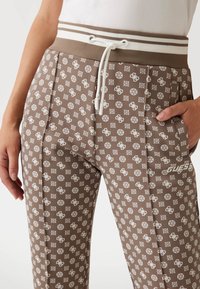 Hnedé jogger nohavice s vzorom, vyrobené z mäkkej látky, s bielym šnúrkovým pásom a logom. Obsahujú bočné vrecká a dekoračné akcenty.