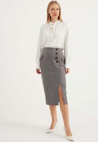Grauer Nadelstreifenrock mit vorderem Schlitz, ausgestattet mit schwarzen Knöpfen und aufgesetzten Taschen. Kombiniert mit einer weißen Bluse und weißen High Heels.
