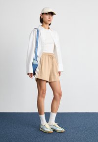 Beige shorts, hvit tank top, oversized hvit knapper skjorte, kremfarget caps, blå crossover veske, og lyseblå sneakers med hvite detaljer.