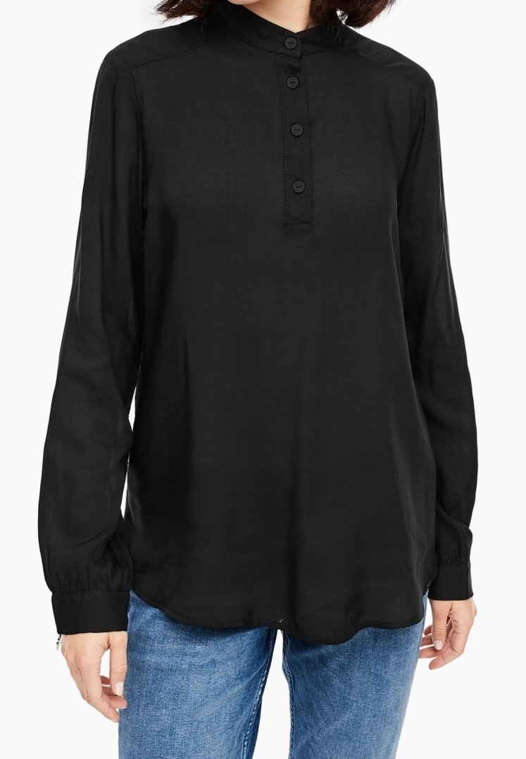 Femme portant un chemisier noir à manches longues avec une patte de boutonnage et un jean bleu, debout sur un fond blanc.