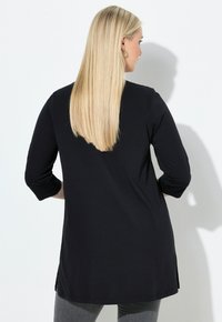 Svart långärmad topp med löst snitt, rundad fåll, sidofack och en slät textur; bärs över grå byxor, sedd bakifrån.