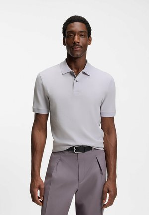 Mężczyzna ubrany w jasnoszarą, krótkorękawową koszulkę polo włożoną w marszczone spodnie w kolorze mauve, z czarnym paskiem, stojący na białym tle.