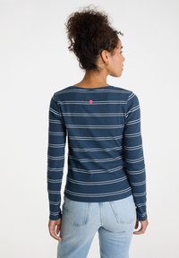 Langärmliges, dunkelblaues Rippenhemd mit weißen horizontalen Streifen. Verfügt über einen kleinen rosa Akzent am hinteren Halsauschnitt. Kombiniert mit hellblauen Jeans.