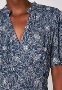 Gros plan sur une robe bleu marine avec des motifs floraux blancs complexes, un décolleté en V froncé, des manches courtes et une taille élastiquée.