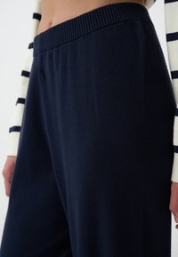 Pantalons en tricot bleu marine avec une taille côtelée, présentant une texture lisse et une coupe ample. Le tissu montre peu de plis et a un aspect uniforme.
