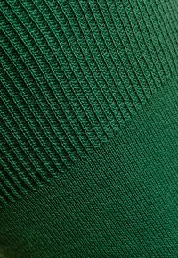 Nahaufnahme von grünem Stoff, der eine gestrickte Textur mit vertikalem Rippenmuster zeigt, das in eine glatte Strickoberfläche übergeht.