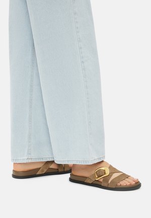 Brązowe zamszowe klapki z szerokim paskiem i złotym akcentem w postaci klamry, zestawione z jasnoniebieskimi szerokimi spodniami.