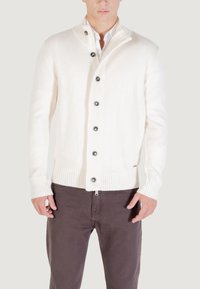 Hombre vestido con un cárdigan blanco de botones sobre una camisa blanca y pantalones marrón oscuro, de pie contra un fondo claro y liso.