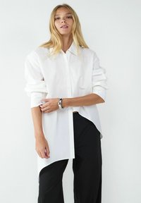 Weißes Hemd mit spitzem Kragen, mit hochgeschlagenen Ärmeln und asymmetrischem Saum, kombiniert mit schwarzen, lockeren Hosen.