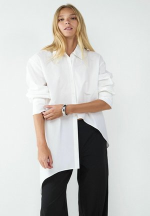 Chemise blanche à boutons avec un col pointu, ornée de manches roulées et d'un ourlet asymétrique, associée à un pantalon noir ample.