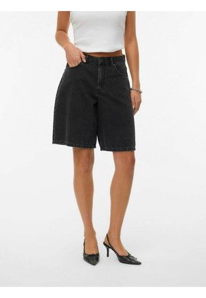 Kvinne har på seg svarte høyhusta vide shorts i denim, hvit croppet topp, svarte slingback hæler og sølvarmbånd, står mot hvit bakgrunn.