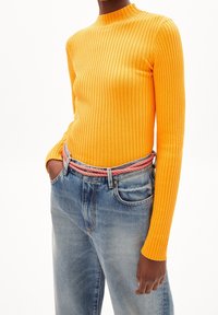 Haut jaune à manches longues côtelé, associé à un jean bleu clair. Le jean a une coupe décontractée et une ceinture rouge et blanche à motif.