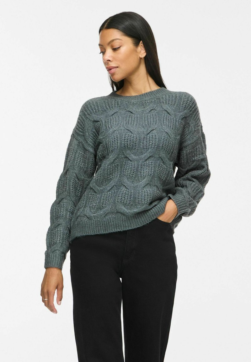 Femme aux longs cheveux foncés portant un pull en maille torsadée gris et un pantalon noir, debout avec une main dans la poche, regardant vers le bas sur le côté.