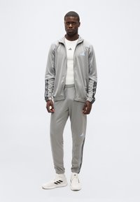 Άνδρας που φοράει γκρι φόρμα Adidas με μαύρες ρίγες και λευκά παπούτσια Adidas, στέκεται μπροστά σε απλό φωτεινό φόντο.