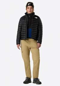 Sort dunjakke over en blå fleece, beige cargobukser og mørke sko. Personen har på seg en camo-lue og jakken har en glidelåsdetalj.