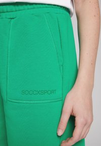 I pantaloni della tuta verdi in cotone presentano una tasca frontale e la scritta "SOCCXSPORT" stampata vicino all'orlo. Il tessuto ha una texture liscia e una vita elasticizzata.