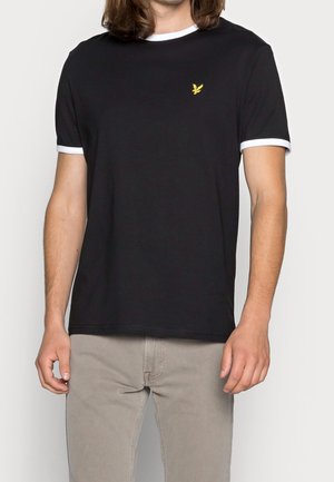 Mann trägt ein schwarzes T-Shirt mit weißen Bündchen an Ärmeln und Kragen sowie einem kleinen gelben Vogel-Logo auf der Brust, kombiniert mit beigen Hosen.